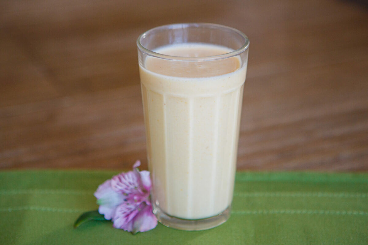 Healthy Shot peach smoothie in tall glass on top of a green tablecloth with a purple flower